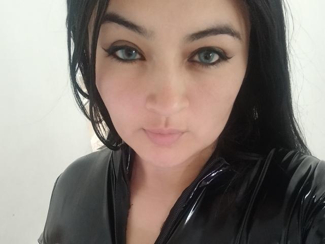 LindaSophieX with black hair, green eyes, and black latex outfit, showing a slight pout. Her makeup includes winged eyeliner. Her tits are not visible in this shot.