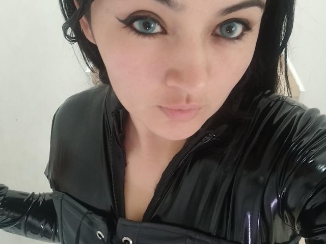 LindaSophieX with big blue eyes, wearing a shiny black latex top, looking up at the camera with pouty pink lips. Her black hair is slightly messy.