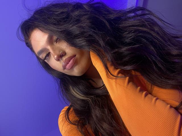 BabeGigi with long, wavy black hair, wearing an orange ribbed shirt, nose piercing, and purple-blue background, looking down with a slight smile, full lips, and subtle makeup.