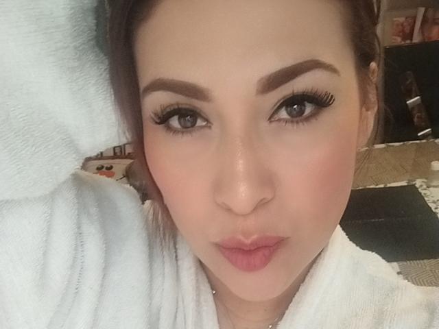 SandiaHot's face with pouty pink lips, thick brown eyebrows, long black eyelashes, and brown eyes, wearing a white towel, looking straight at the camera.