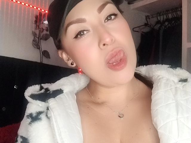 SandiaHot with pouty lips, wearing a black baseball cap, white quilted jacket, and a heart necklace, sticking out her tongue. She has dark eyebrows, red earrings, and a nose ring.