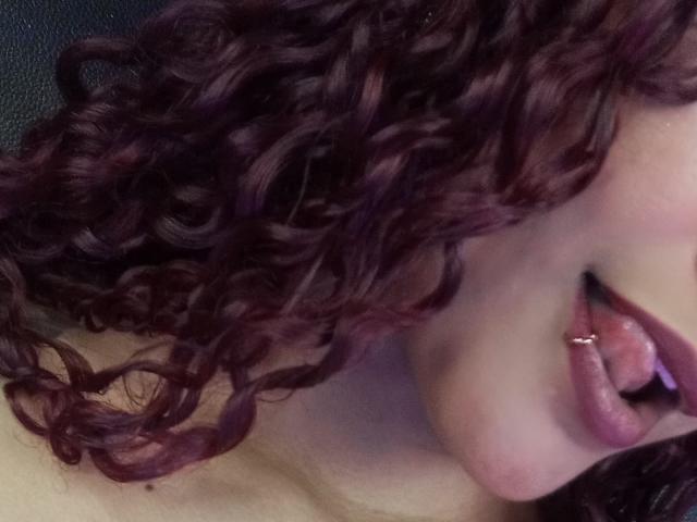 A selfie of CumLovingSquirter with curly purple hair, tongue out, showing a silver lip piercing, and slightly open pinkish lips. CumLovingSquirter's face is partially visible.
