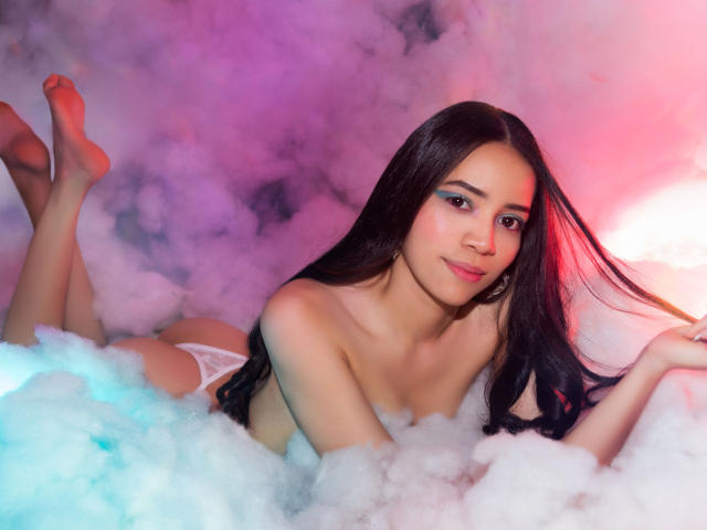 KendallParker with long black hair, lying in fluffy white smoke, wearing white lace panties, showing her tits and ass, smiling with red and pink light on her face.