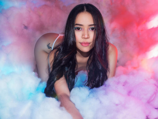 KendallParker with long black hair, looking at camera, on knees in pink and blue fluffy smoke, wearing white thong, showing ass, small tattoo on right butt cheek.