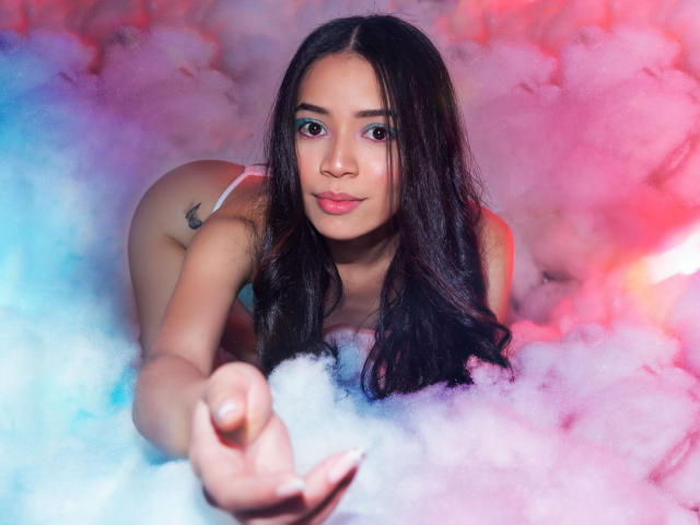 KendallParker with long dark hair, blue eyeshadow, and pink lipstick, on her knees in white underwear, reaching forward, surrounded by pink and blue smoke. Boobs visible, ass up.