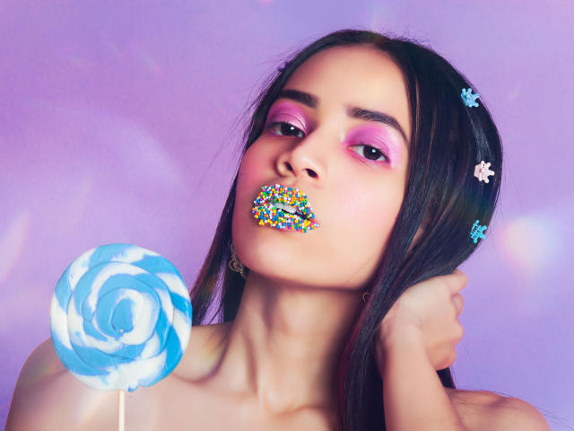 Kendall Parker mit rosa Lidschatten, bunter Lippenstift und einem blauen, gespiralten Bonbon in der Hand, hält ein blaues, gespiraltes Bonbon vor einem violetten Hintergrund. Ihre Brüste sind teilweise sichtbar..