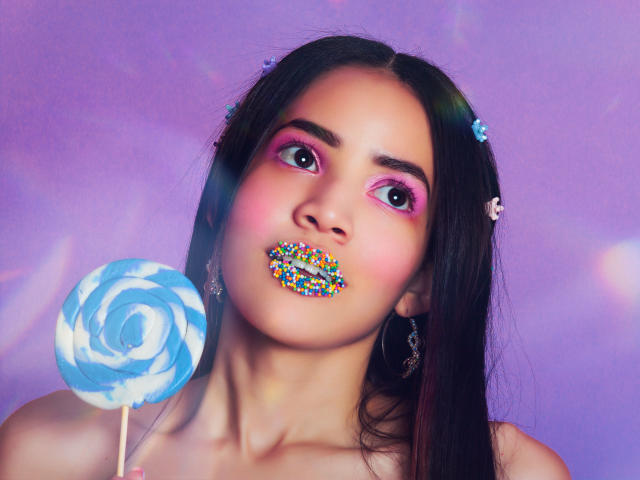 KendallParker has dark hair, pink eyeshadow, colorful lip glitter, holds blue swirl lollipop, wearing earrings, against purple background, looking up with wide eyes.