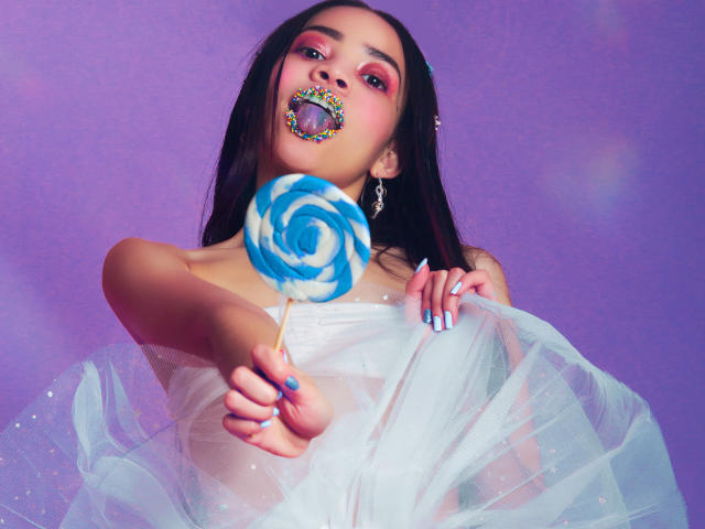 KendallParker with long black hair, holding a blue and white swirl lollipop, wearing a white dress with a fluffy skirt, and colorful lipstick, against a purple background.
