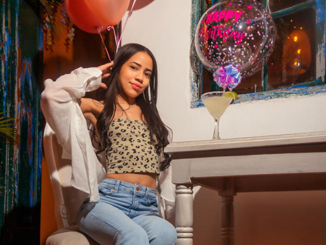 KendallParker with long black hair, wearing a leopard-print crop top and blue jeans, sitting at a table with a drink, balloons, and a "Happy Birthday" balloon.