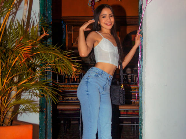 KendallParker with long dark hair, white sheer bralette, and blue high-waisted jeans, smiling, hand in hair, standing in doorway, holding black purse, small butt, slim, cute.