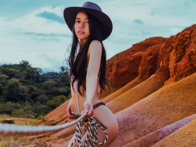 KendallParker with long black hair, wearing a black hat, pink bikini, and holding a rope, standing in front of red rock hills, showing her small ass and cute face.