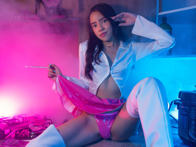 KendallParker with long dark hair, wearing a white open shirt, pink shiny panties, and white thigh-high boots, holding a wand, sitting with legs spread, under pink and blue lights.