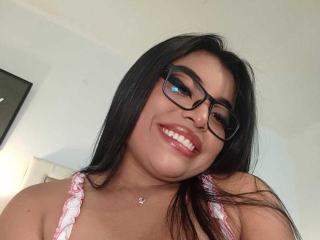 AlexaMadisson has dark, straight hair, wears black glasses, and smiles with pink lipstick, showing white teeth. She's in a pink and white top, and has a small necklace.