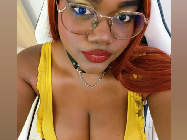 RoseBeckers with red hair, wearing glasses, yellow dress, and black choker with chain, has big tits and red lipstick, looking at camera.