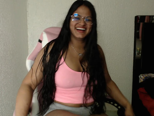 SweetAlma69, Latina with long black hair, glasses, pink tank top showing her tits, smiling, sitting on a pink-and-white chair, wearing grey shorts, in a dimly lit room.