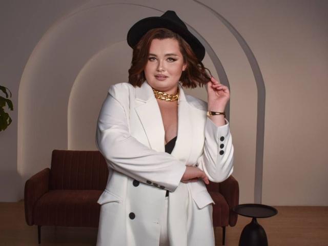 JessicaJoye con un camice bianco, un cappello nero, una collana dorata e un braccialetto, che tiene il cappello con una mano, mostrando il seno in un reggiseno nero, in piedi di fronte a un divano..