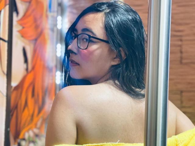 JewelryQueen, com cabelo escuro, óculos e bochechas rosadas, olhando por cima do ombro, sem blusa, com uma toalha amarela, perto de um poste de metal, com uma obra de arte na parede em tons de laranja e preto ao fundo..
