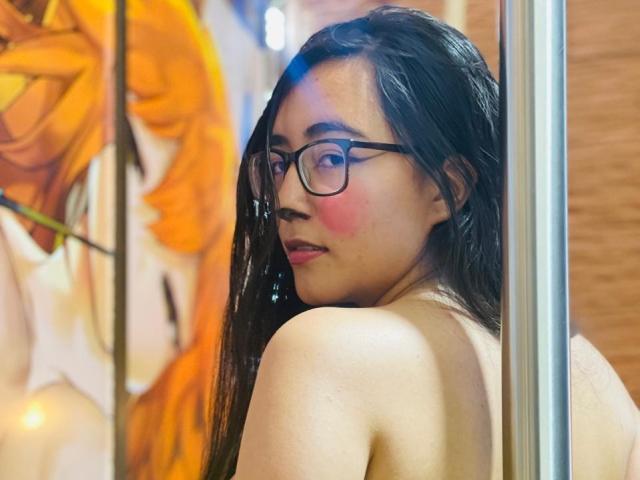Uma mulher seminua, chamada JewelryQueen, usando óculos, com bochecha rosada, cabelo preto longo e costas nuas, olhando por cima do ombro, ao lado de um mastro. Seus seios estão parcialmente visíveis. O fundo tem arte colorida e borrada..