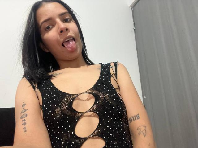 KaylinJulithe with black hair, tongue out, wearing a black, cut-out, polka-dot top, showing tattoos on arms, in a selfie. She has small tits and a cute face.