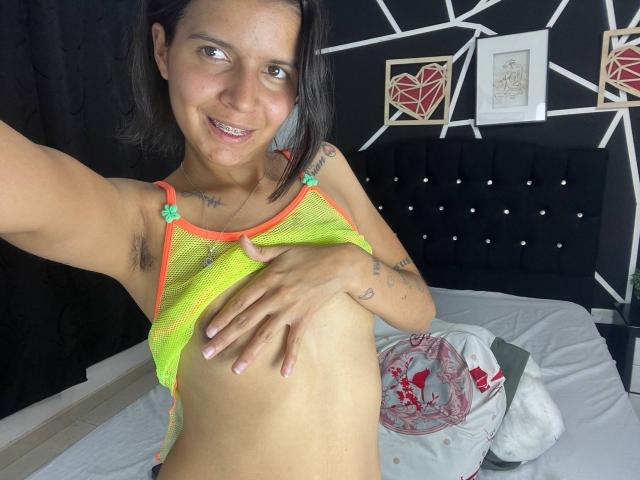 KaylinJulithe with small tits, armpit hair, smiling, wearing neon green mesh top, hand on chest, black headboard, tattoo on neck, bed with white and red pillows, dark wall.