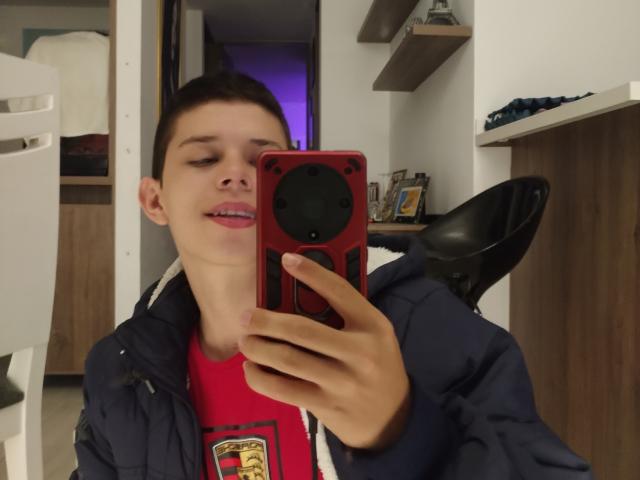 JhoanVelezX con cabello castaño corto, chaqueta azul, camisa roja, sosteniendo un teléfono rojo con cámara negra, sonriendo con la lengua afuera, tomando un selfie en una habitación..