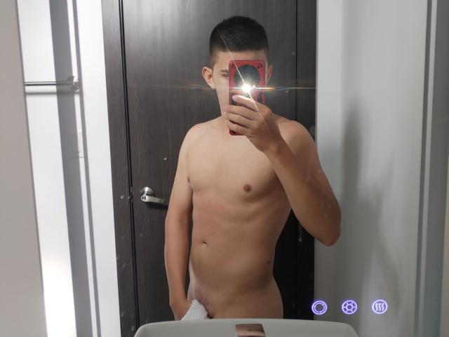 Naked JhoanVelezX with a towel covering his flaccid, uncircumcised dick, holding a red phone in a mirror selfie, showing a shiny reflection and a slim, toned body.