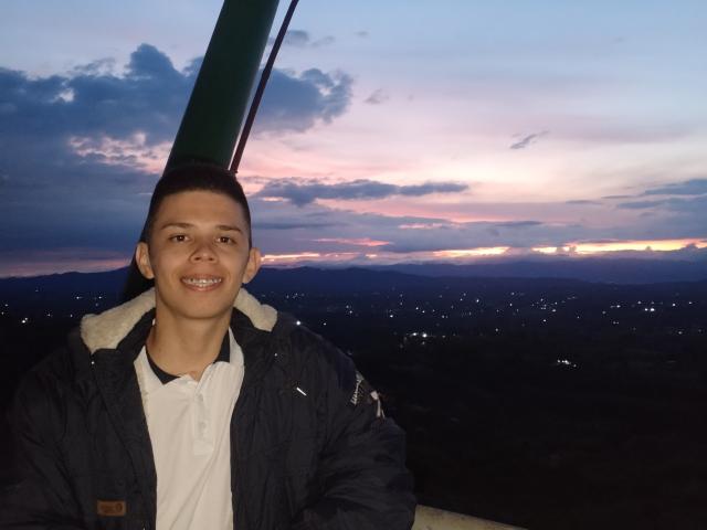 JhoanVelezX, avec des cheveux bruns courts, souriant, portant une veste sombre par-dessus un polo blanc, debout sur un siège de télésiège vert au crépuscule, avec un ciel violet et rose en arrière-plan..