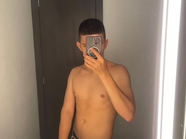 JhoanVelezX, sin camisa, con un pecho claro, liso y de piel clara, sosteniendo un teléfono que le cubre la cara, reflejo en un espejo, cabello castaño, pezones pequeños, de pie frente a una luz brillante..
