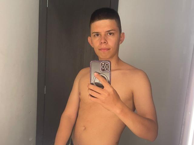 JhoanVelezX, torse nu, avec une courte coupe de cheveux, tenant un téléphone devant son visage, regardant la caméra, souriant légèrement, montrant une partie supérieure du corps lisse et tonique..