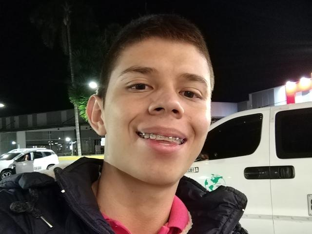 JhoanVelezX, avec des cheveux bruns courts, souriant, montrant des bagues orthodontiques en métal, portant une veste sombre par-dessus une chemise rose, devant une ambulance blanche la nuit..