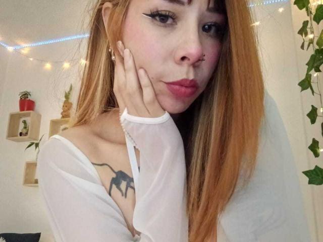AriiannaHot with long orange hair, white sheer top, black spider tattoo on left shoulder, pouty lips, nose ring, and hand on cheek, looking at camera.