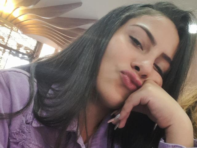 Ruby Jade, avec ses longs cheveux noirs, son nez légèrement retroussé et ses lèvres roses pulpeuses, reposant son menton sur sa main, portant une chemise violette, regardant de manière séduisante sur le côté..