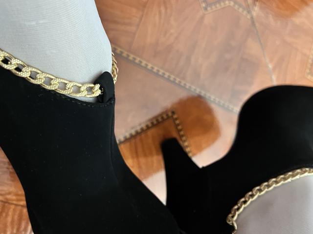 ShantalSummer's black high-heeled boot with gold chain detail and white sock, showing the heel and ankle, on a wooden floor. Boot is positioned to the left, heel to the right.