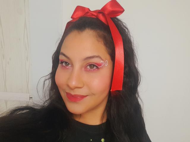 RubyJades avec des cheveux noirs ondulés, un ruban rouge dans ses cheveux, un eye-liner rose, un cœur en eye-liner blanc, du rouge à lèvres rouge, souriante, portant un haut noir, fond blanc, lumière douce..