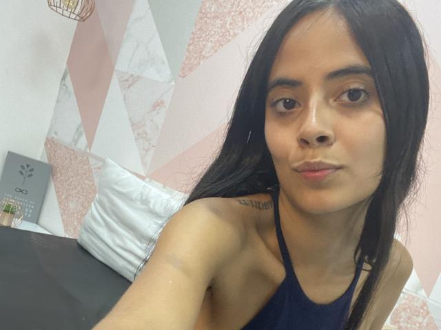 TamaraPeach has long black hair, brown eyes, and a small tattoo on her left shoulder. She's wearing a dark blue halter top, showing her small tits and pouty lips.