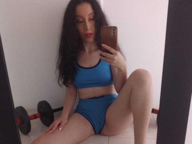 KendraSimons with long black hair, blue sports bra, and matching shorts, sitting on the floor, legs spread, holding a phone, with dumbbells in the background.