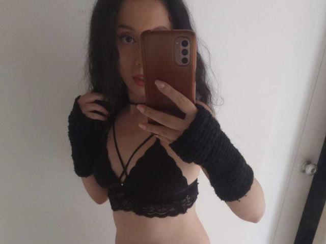 KendraSimons con un sujetador de encaje negro, guantes negros sin dedos y cabello negro largo, sosteniendo un teléfono, mostrando sus pechos y abdomen en un selfie frente al espejo..