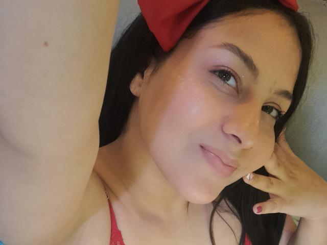 RubyJades with long black hair, red bow, and red bra, smiling slightly, showing her nose, eyes, and pinkish skin. Her right hand touches her cheek.