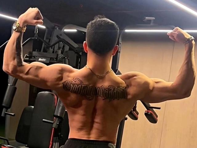 InkAngelo's muscular back with a tattoo reading "Strongest" across his shoulders, flexing his arms in a gym, showcasing defined muscles and a gold wristwatch.