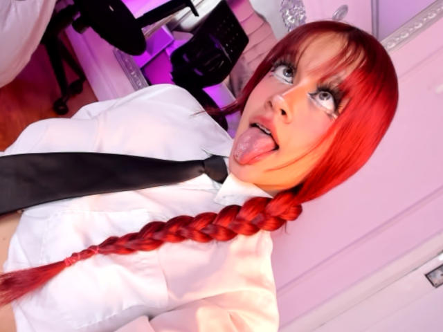 Ada Ferreira mit roten Zöpfen, weißem Hemd, schwarzem Krawatten und schwarzem Augen-Make-up, die die Zunge herausstreckt, hat blaue Augen und blickt nach oben. Ihre Brüste sind klein..