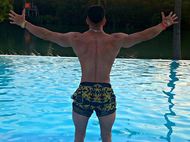 InkAngelo's muscular back, arms outstretched, wearing yellow and black swim trunks, a silver chain, and red wristband, standing in a blue pool. InkAngelo's butt is visible.