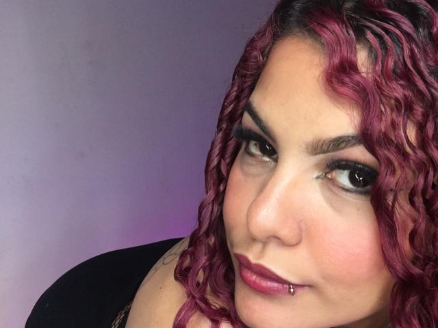CumLovingSquirter with pink curly hair, brown eyes, dark eyeliner, pink lipstick, nose piercing, smiling, looking at camera, wearing black top, purple background, slight pink light.