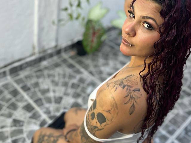 Curly red-haired CumLovingSquirter with tattoos, nose ring, and brown eyes, looking back, wearing a white tank top, showing a tattooed shoulder and black shorts.