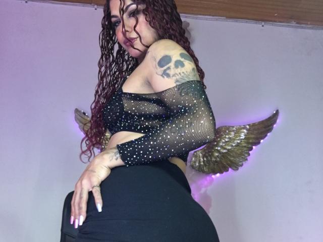 CumLovingSquirter with curly brown hair, skull tattoo, black mesh top, and black pants, showing her ass, wearing gold winged accessory, in a low angle shot.