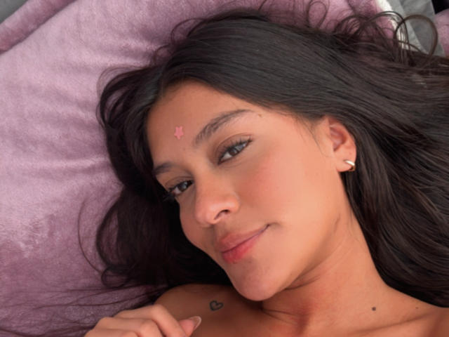 ThaliaAres lying on a purple pillow, smiling, with dark brown hair, a small pink star on her forehead, a heart tattoo on her shoulder, and small ear piercings.