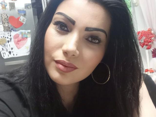 BeliseChappelleX-hot has dark, wavy hair, thick eyebrows, long eyelashes, and pink lipstick. She's wearing a black top and a gold hoop earring, looking slightly to the side.