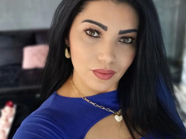 BeliseChappelleX-hot with long black hair, thick eyebrows, and pink lipstick, wearing a blue top with cutouts, gold necklace, and earrings, staring directly at the camera.