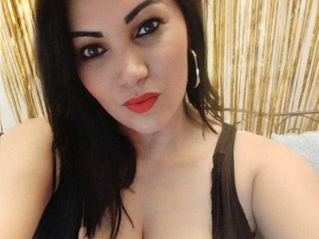BeliseChappelleX-hot with dark hair, red lipstick, hoop earrings, and a black top showing cleavage, looking at the camera with a slight smile. Boobs are visible.