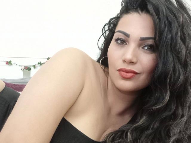 BeliseChappelleX-hot with wavy black hair, red lipstick, and hoop earrings, wearing a black strapless top, showing her bare shoulders and upper chest.