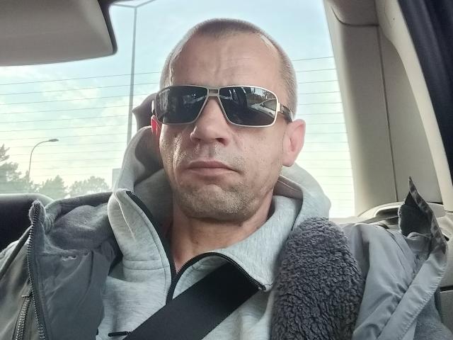 GejPolska with sunglasses, a grey hoodie, and a black seatbelt, looking serious, sitting in a car with a grey jacket and a fuzzy shoulder strap.